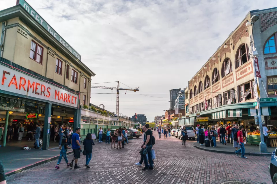 View of stone-paved street with pedestrians and "Farmers Market" neon sign on left and old buildings on right in Seattle, Washington